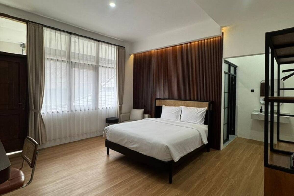 A modern bedroom with a large bed, a wooden floor, a window with sheer curtains, and a wooden headboard.