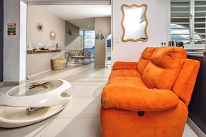 Ein modernes Wohnzimmer mit einem leuchtend orangefarbenen Sofa, einem Glastisch und Meerblick durch große Fenster.