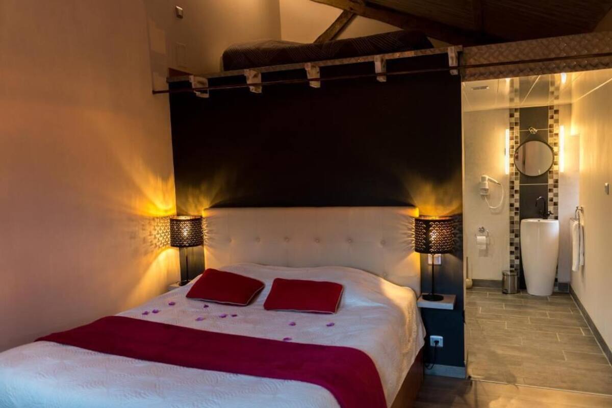 A bedroom with a large bed, two bedside lamps, a red throw blanket, and a bathroom visible through an open door.