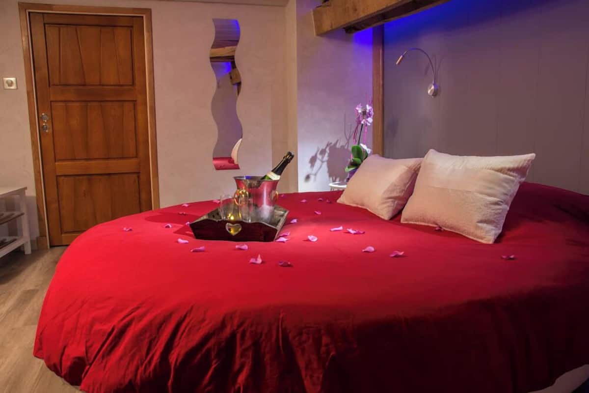 A bedroom with a red bedspread, a wooden door, and a small table with a bottle and flowers.