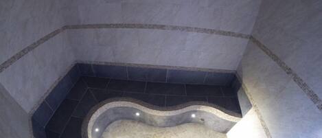 Una bañera grande y curvada con piso de mosaicos, rodeada de baldosas oscuras y un borde alicatado.
