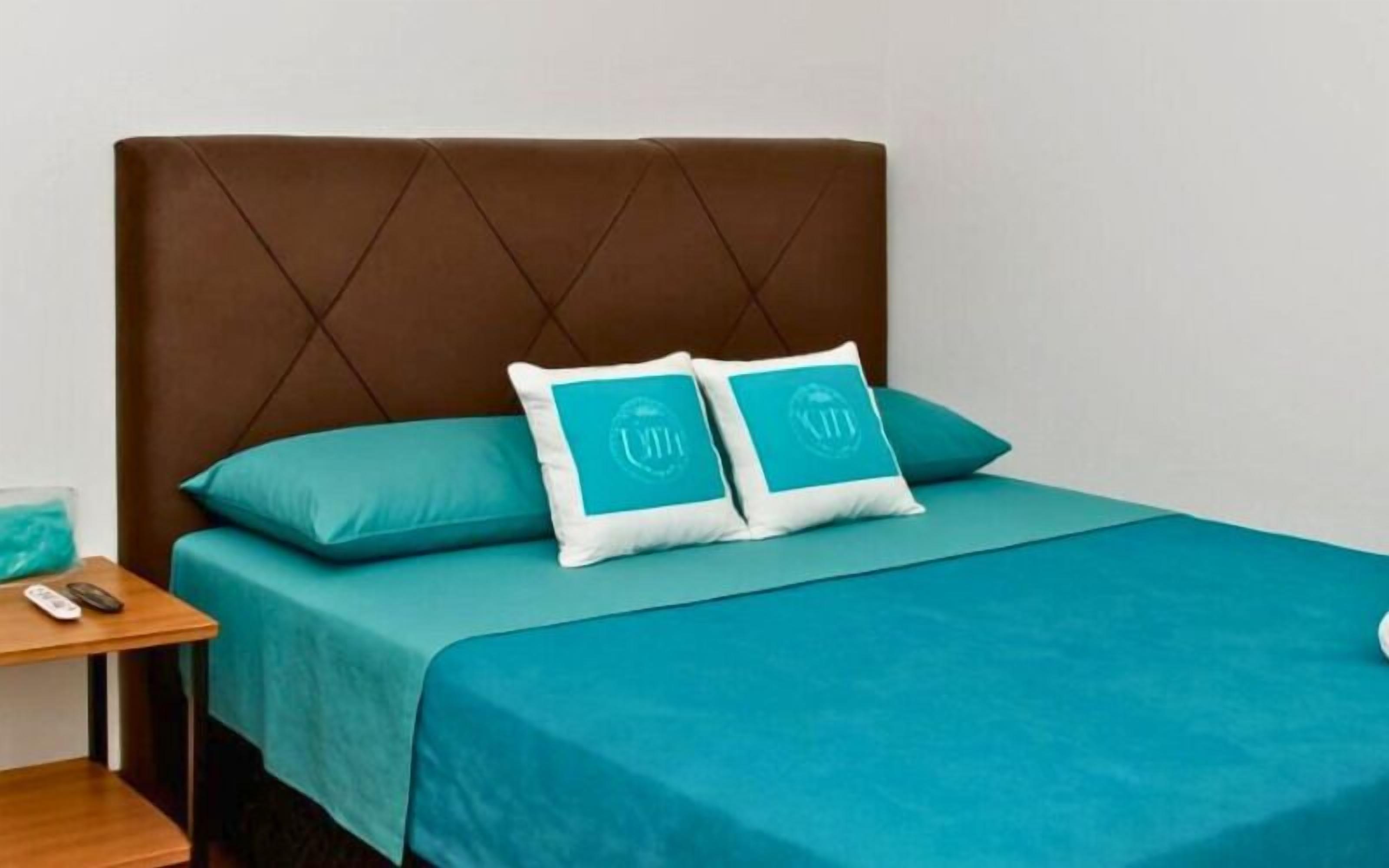 Ein Bett mit braunem Kopfteil, blaue Bettwäsche und zwei blaue Kissen mit einem Logo.