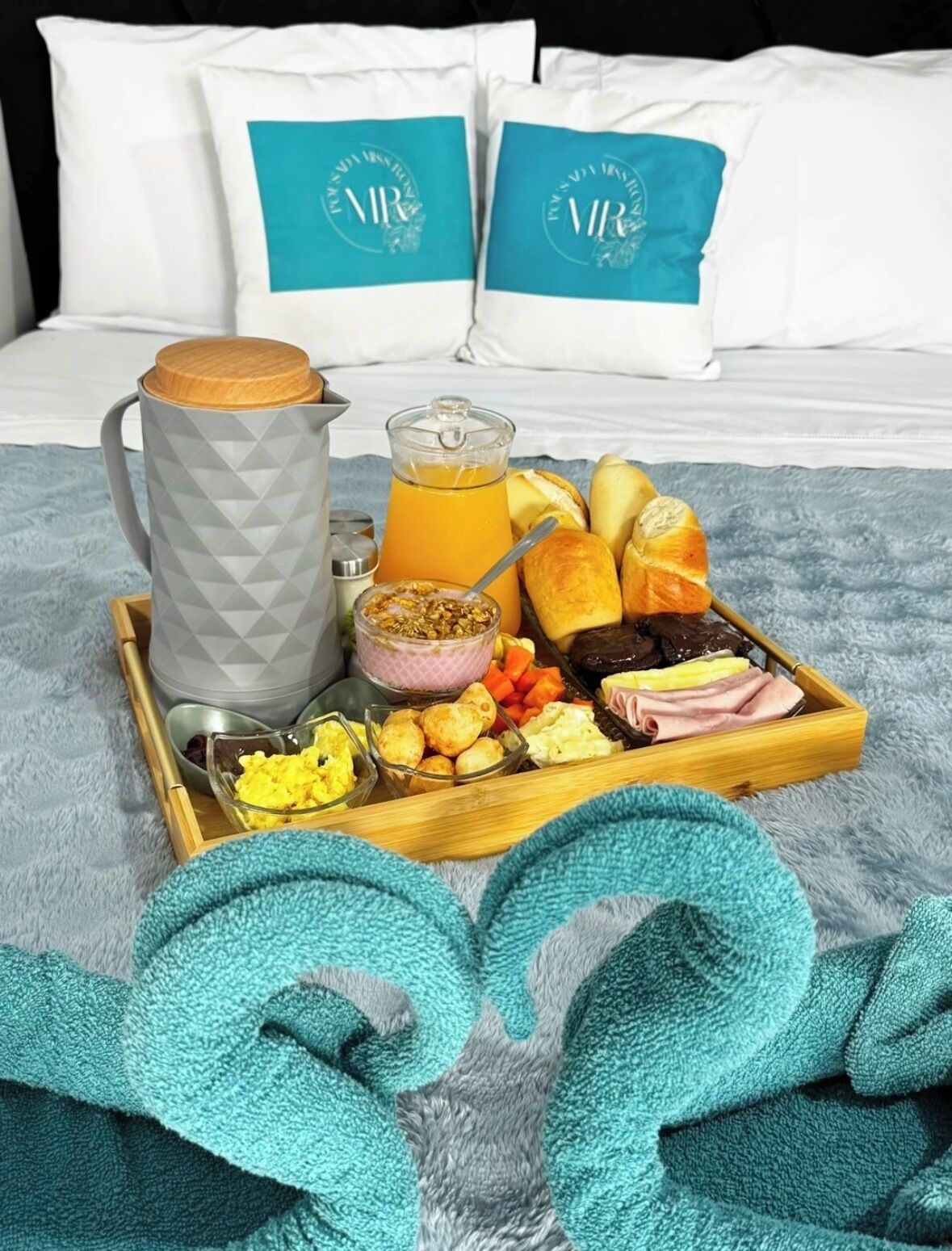 Bandeja de desayuno con huevos, tostadas, jamón y jugo sobre una cama con manta y almohadas azules.