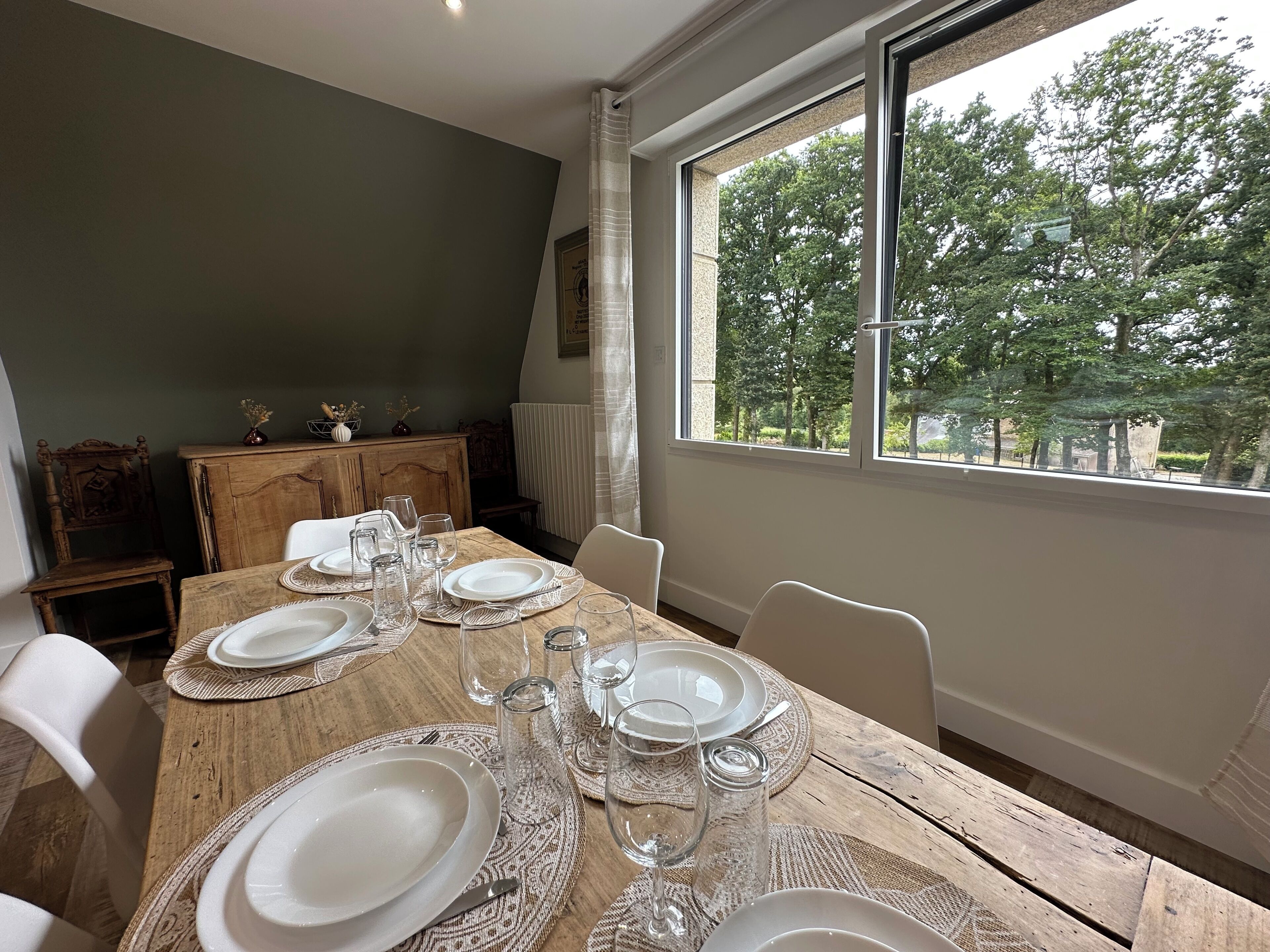 A dining table set with plates, glasses, and cutlery, a wooden cabinet, and a view of trees through a large window.