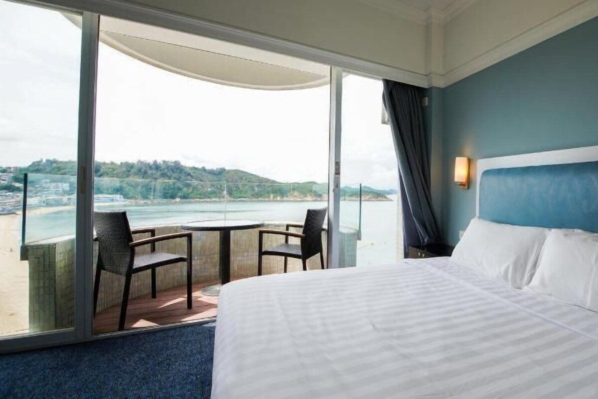 A hotel room with a bed, a view of the beach, and a balcony with chairs.