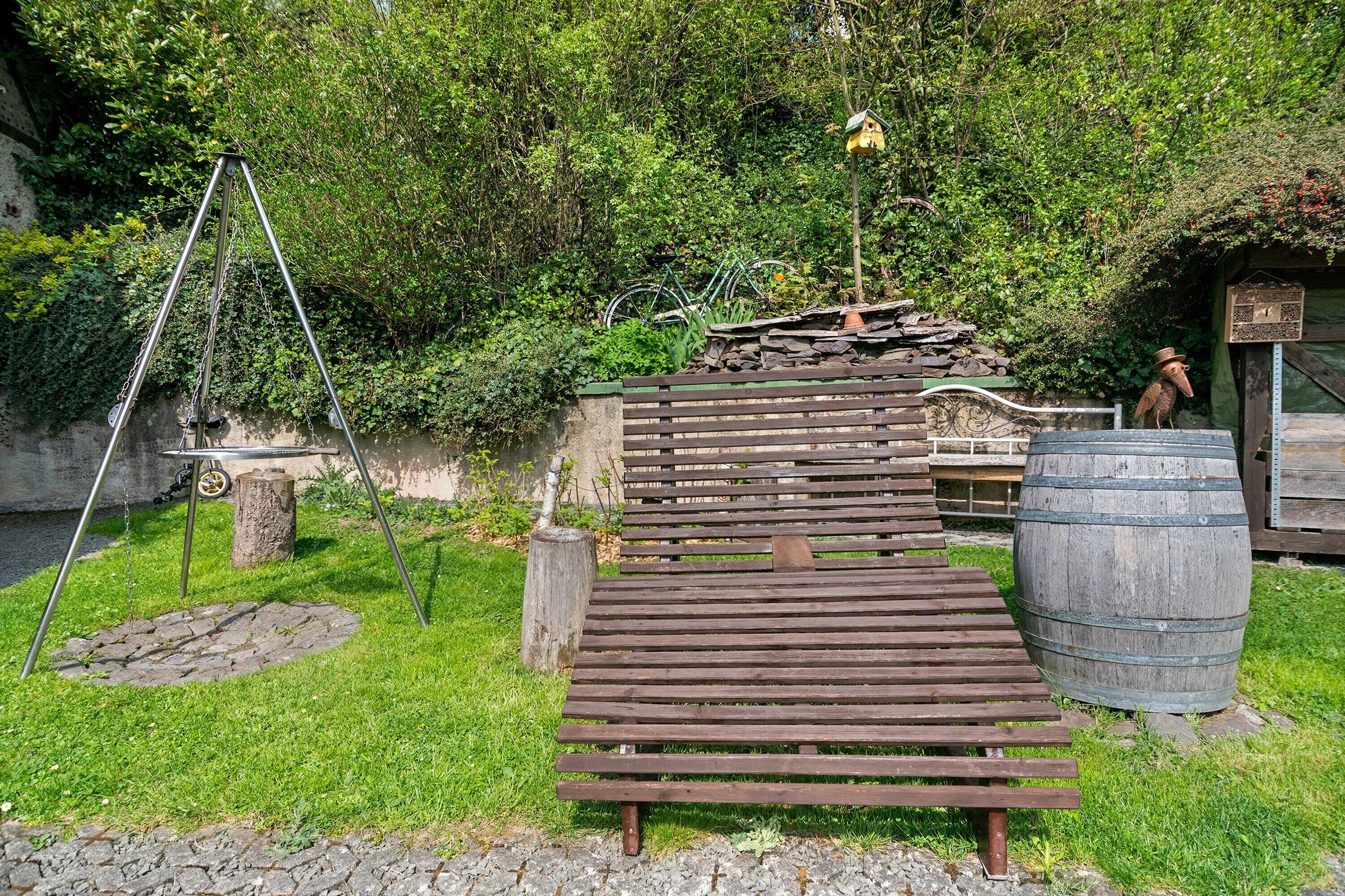 Um banco de madeira, um muro de pedra, um barril de madeira e um bebedouro para pássaros em um jardim.