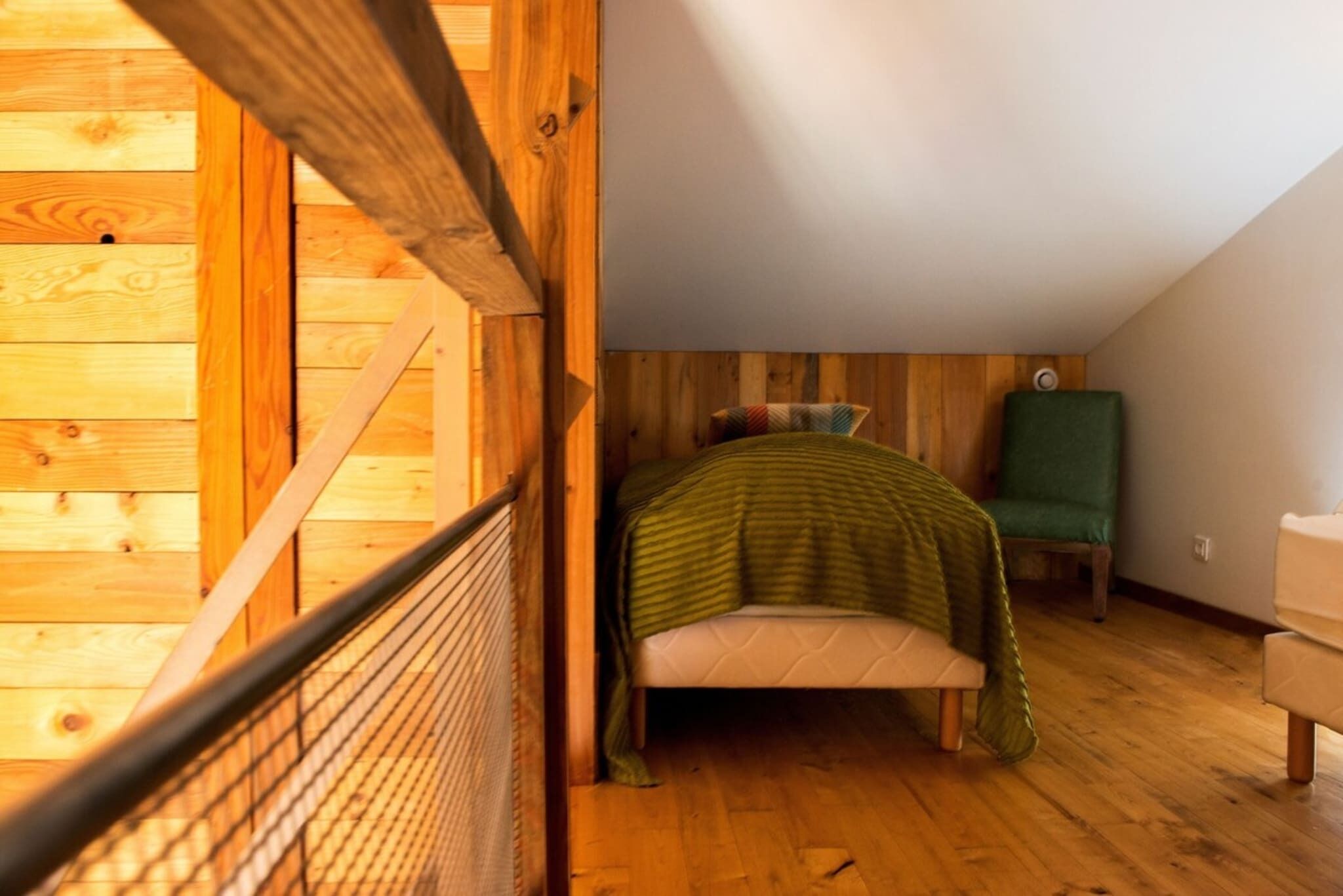 A room with a wooden floor, a bed with a green blanket, a chair, and a wooden wall.