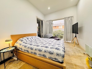 Un dormitorio con cama, televisor sobre un trípode, lámpara y vista al exterior.