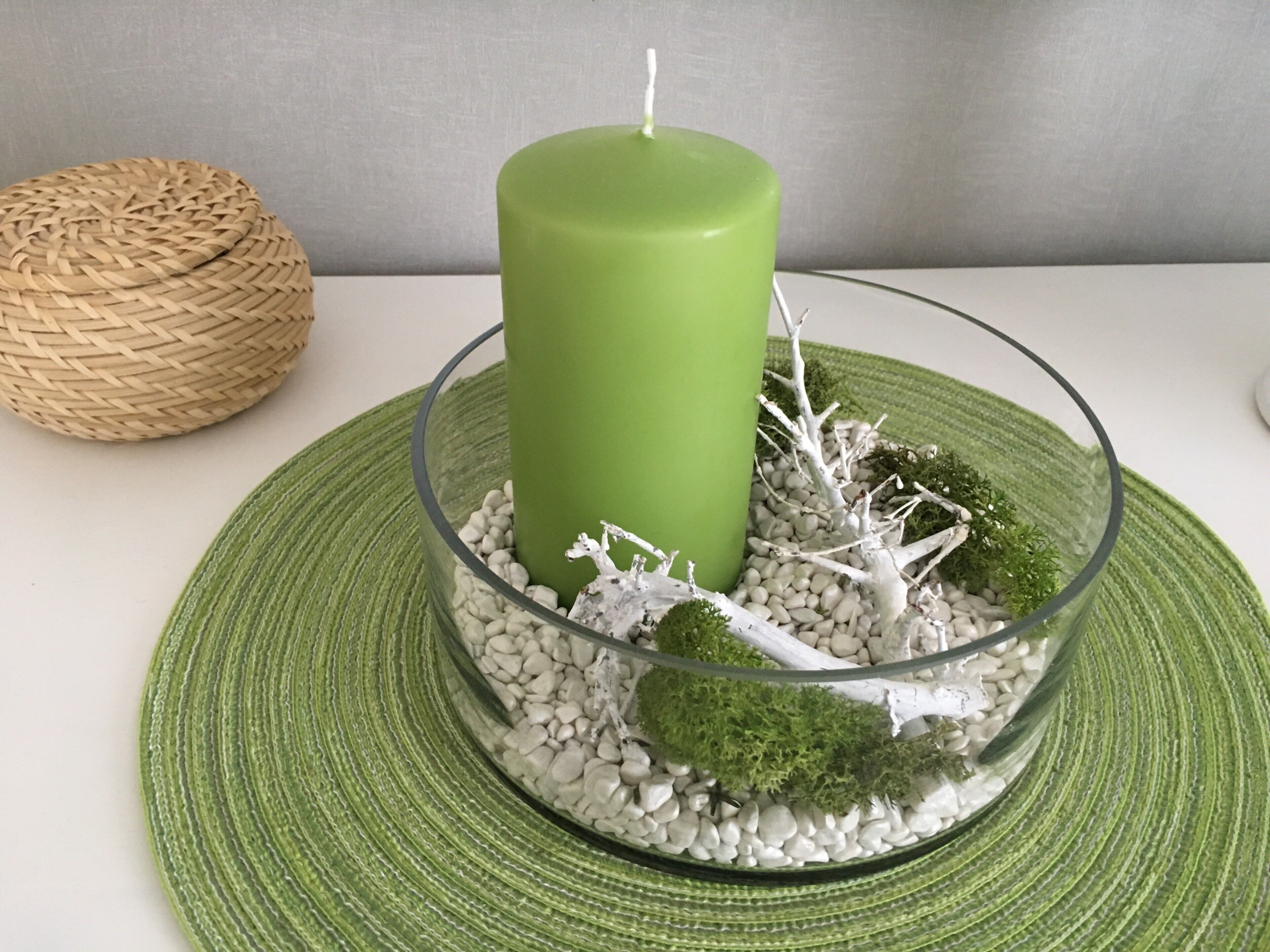 Een groene kaars in een glazen houder met witte kiezelstenen en kleine plantjes, geplaatst op een groene onderzetter.