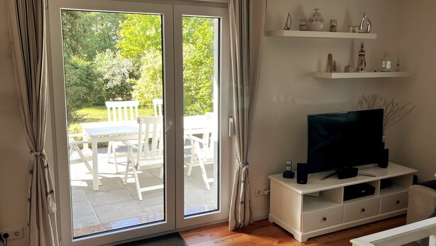 Une pièce dotée d’une porte-fenêtre coulissante donnant sur une terrasse, d’un meuble de rangement blanc abritant un téléviseur, et d’étagères ornées d’objets décoratifs.