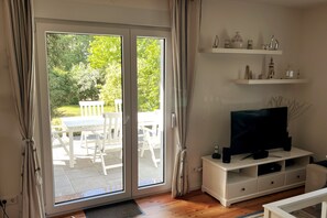 Une pièce dotée d’une porte-fenêtre coulissante donnant sur une terrasse, d’un meuble de rangement blanc abritant un téléviseur, et d’étagères ornées d’objets décoratifs.
