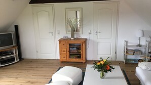 Une pièce avec un canapé blanc, une table blanche ornée d’un vase de fleurs, un meuble en bois rempli de livres et un téléviseur posé sur un meuble.