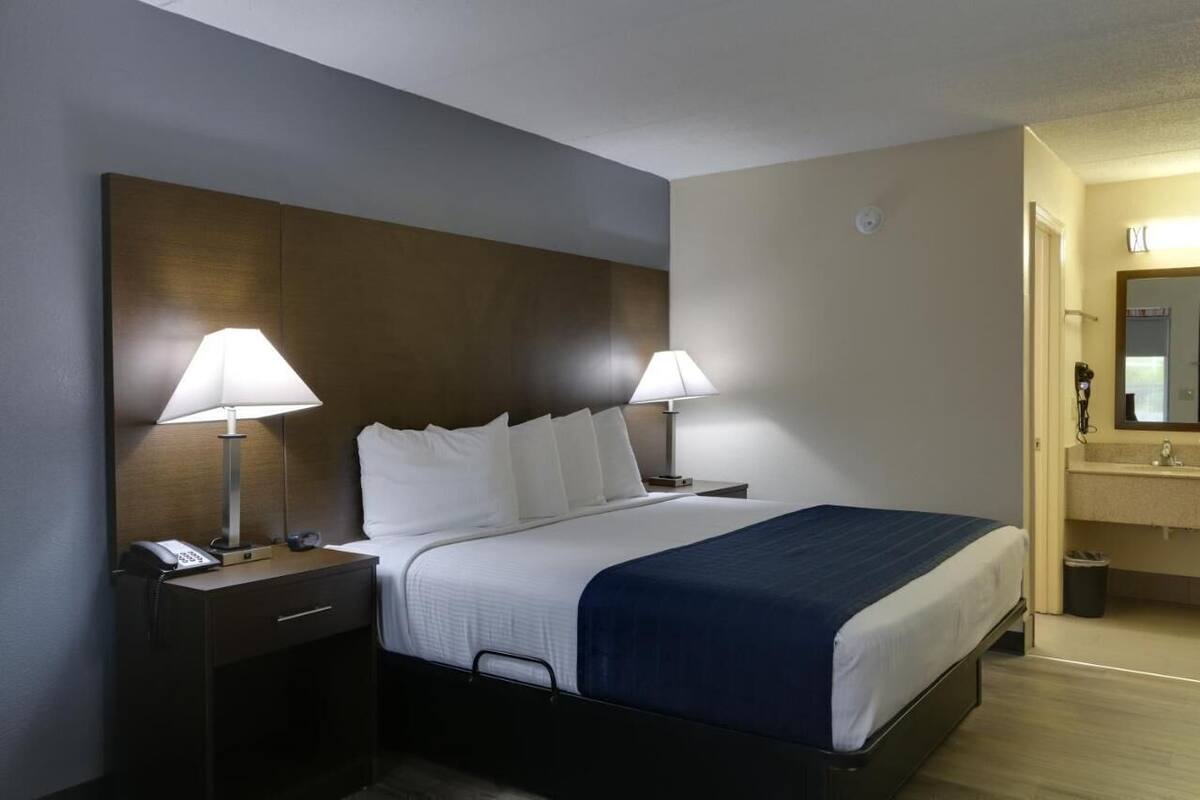 Ein Hotelzimmer mit einem Bett, zwei Nachttischlampen, einem Nachttisch mit Telefon, einer kleinen Mülleimer und einem Badezimmer mit Waschbecken und Spiegel.