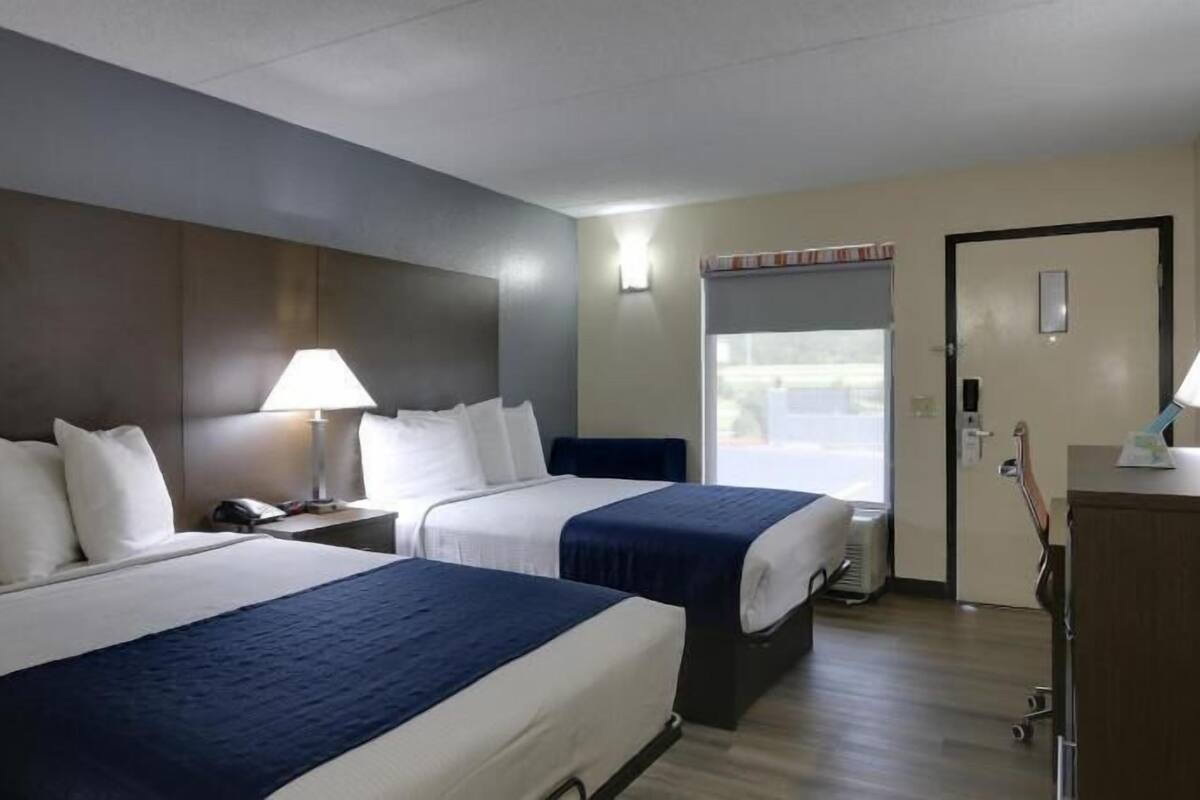 Ein Hotelzimmer mit zwei Betten, einem Schreibtisch, einem Stuhl, einer Lampe und einem Fenster.