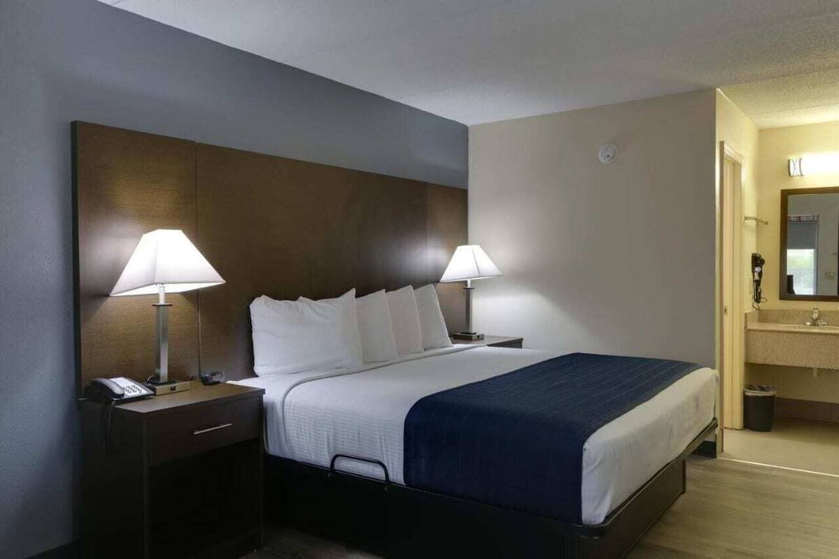 Ein Hotelzimmer mit einem großen Bett, zwei Nachttischlampen, einem Nachttisch mit Telefon sowie einem Badezimmer mit Waschbecken und Spiegel.