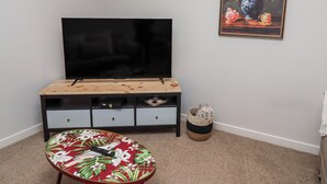 A living room with a TV on a wooden stand, a floral table, a painting, and a woven basket. - Amber Rose (Berwick-upon-Tweed)