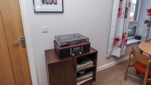 A room with a wooden door, a framed picture on the wall, a radio on a wooden cabinet, a window with floral curtains, and a chair. - Amber Rose (Berwick-upon-Tweed)