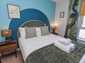 A bed with a woven headboard, two grey pillows, a white duvet, a green throw, and a folded towel. There are two bedside lamps, a small wooden nightstand, and a framed picture on the wall. - Amber Rose (Berwick-upon-Tweed)