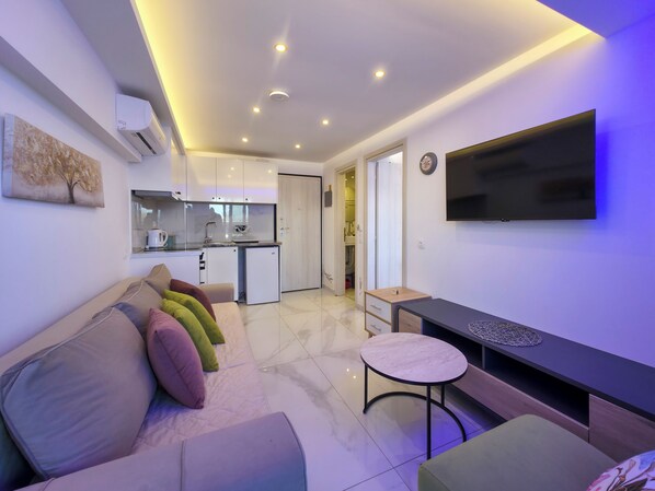 A modern living space with a sofa, a small round table, a flat-screen TV, and a kitchenette.