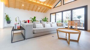 A modern living room with a sofa, coffee table, and chairs, large windows, and wooden ceiling beams. - Le Balzac – Architect-designed home 132m² (Dijon)