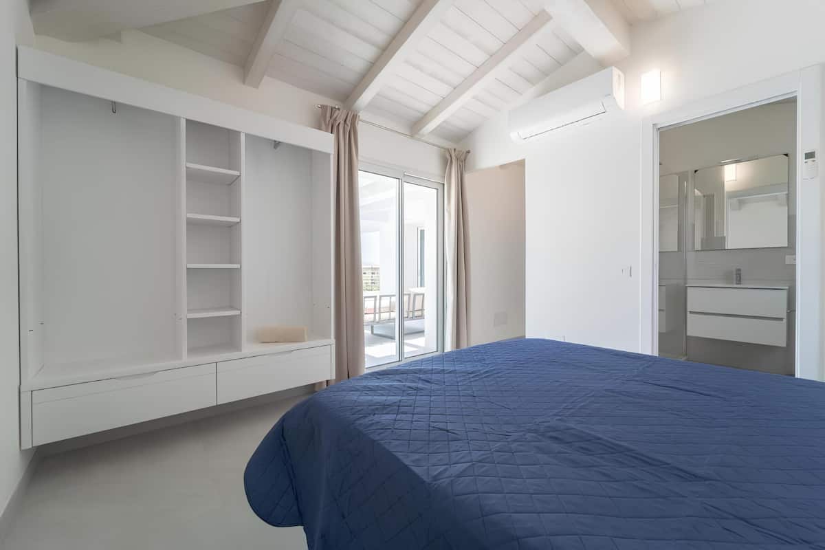 Un dormitorio con una cama grande, un armario, un baño con espejo y una puerta corrediza de cristal a un balcón.