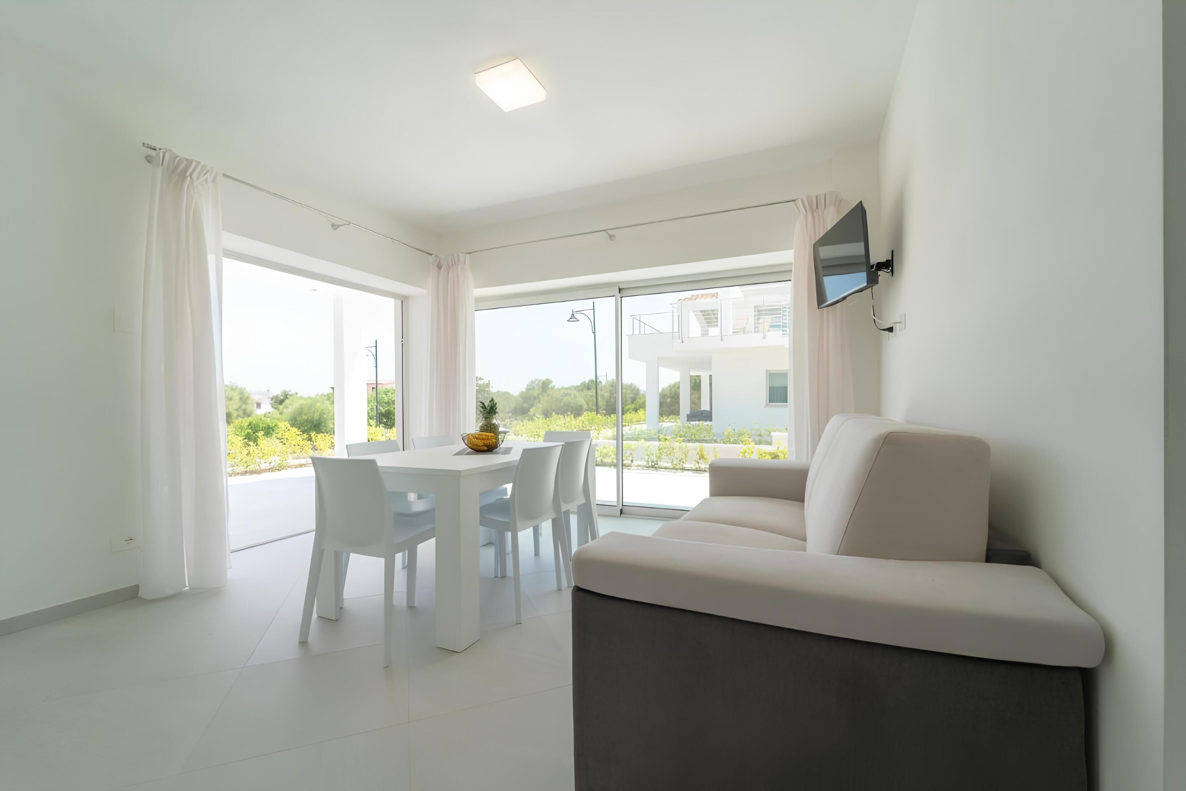 Un salon moderne comprenant un canapé, une table à manger et des chaises, un téléviseur à écran plat fixé au mur et de grandes fenêtres offrant une vue sur la verdure et les bâtiments.