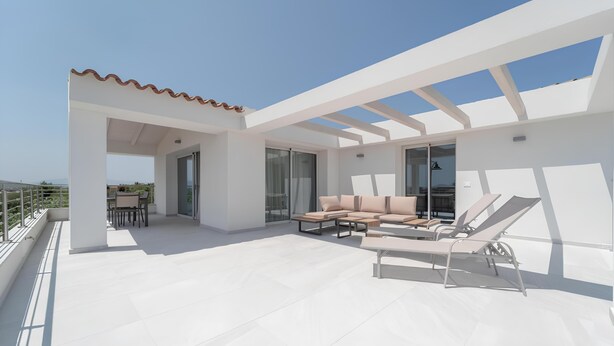 Una terraza moderna en la azotea con suelo de baldosas blancas, un conjunto de sofás color beige y una tumbona.