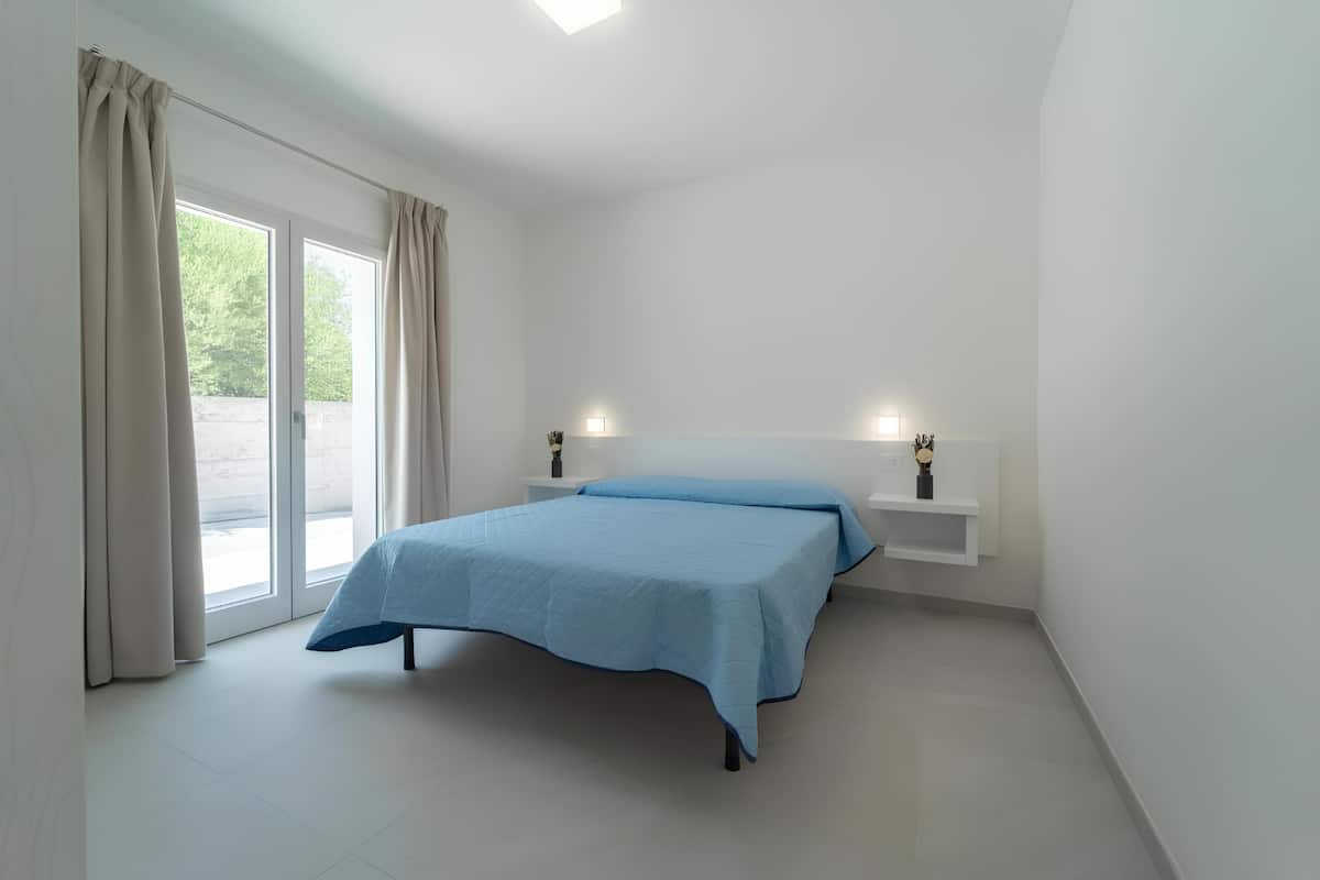 Un dormitorio con cama, cortinas, una mesita de noche, un estante en la pared y una ventana.