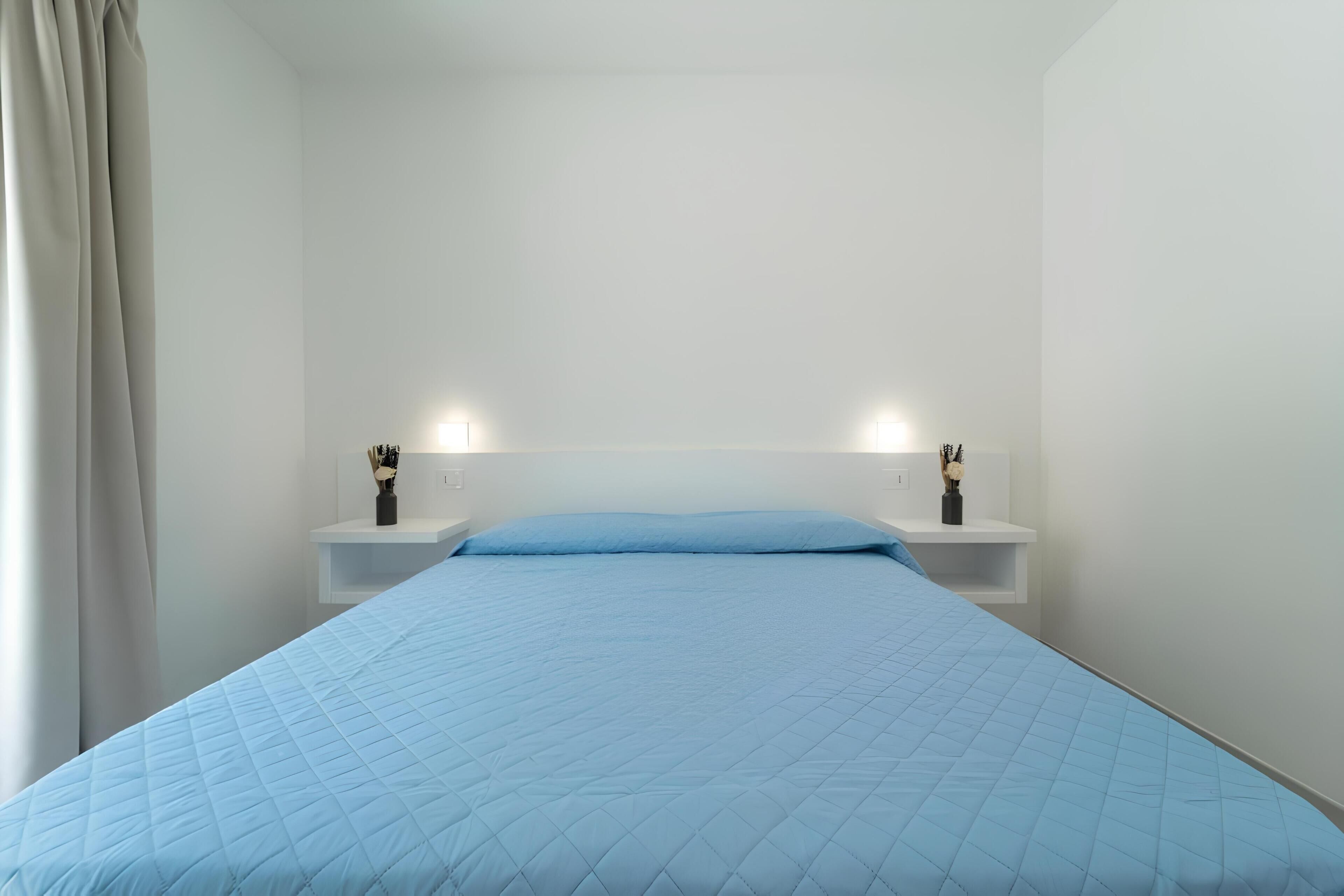 A bed with a quilted blue cover, white walls, and two bedside tables with vases.