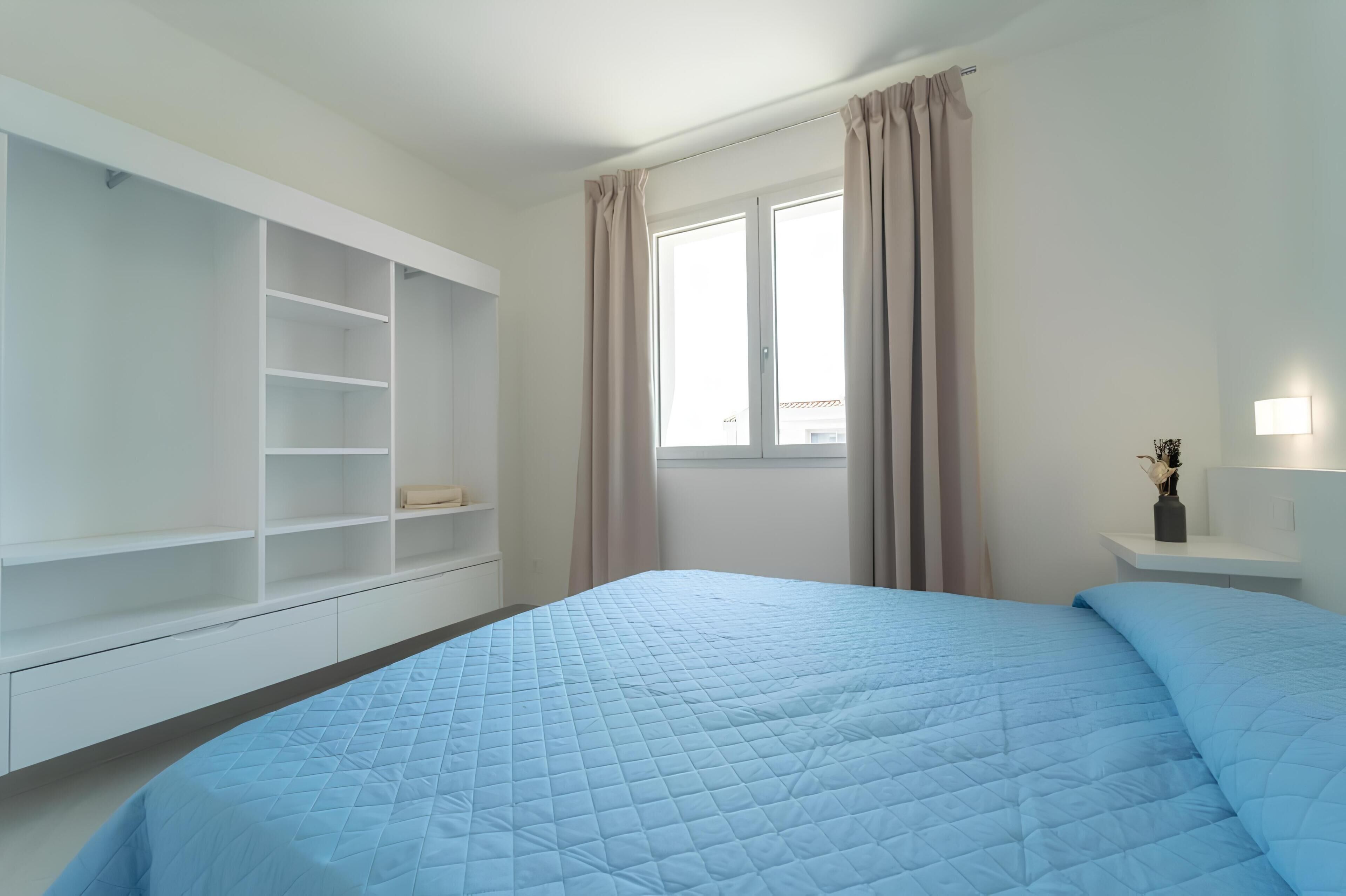 Ein Schlafzimmer mit Bett, Kleiderschrank, einem Fenster mit Vorhängen und einem Nachttisch mit Lampe.