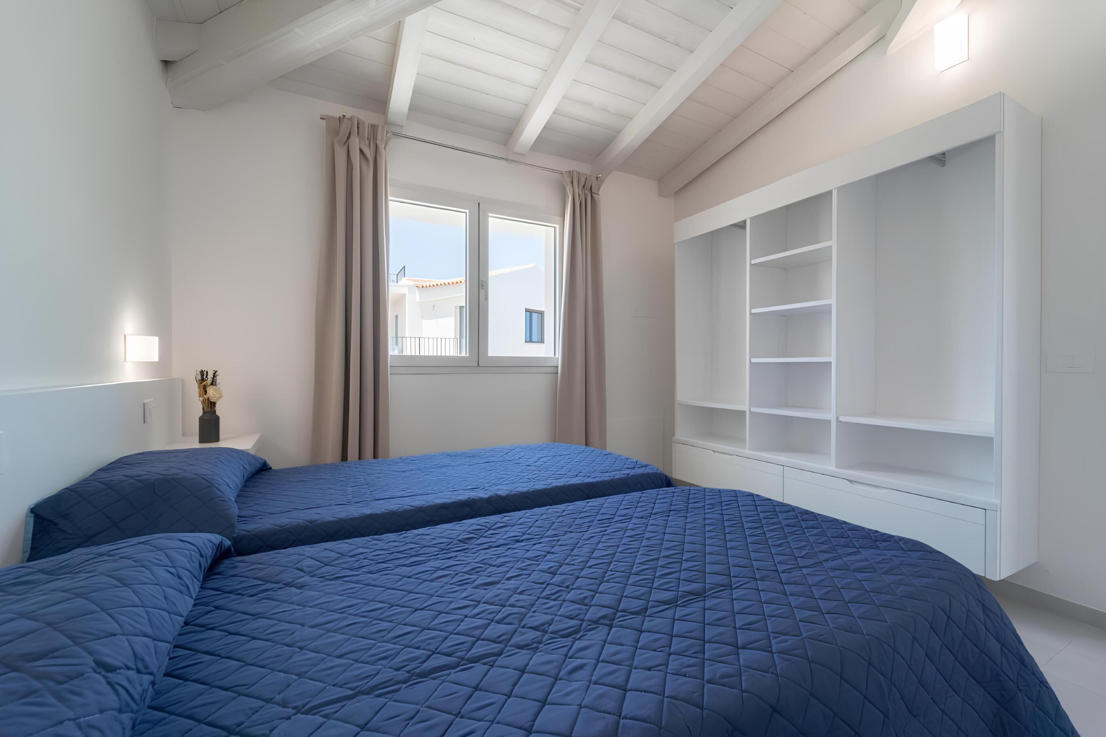 Une chambre avec deux lits, une fenêtre avec des rideaux, une table de chevet avec une lampe et une armoire intégrée.