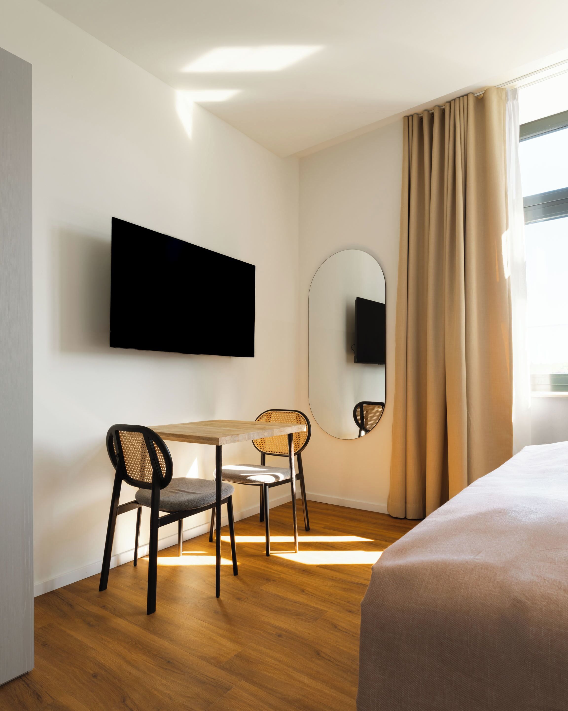 A hotel room with a bed, a desk with two chairs, a flat-screen TV mounted on the wall, a large mirror, and a window with curtains.