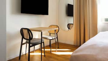 A hotel room with a bed, a desk with two chairs, a flat-screen TV mounted on the wall, a large mirror, and a window with curtains.