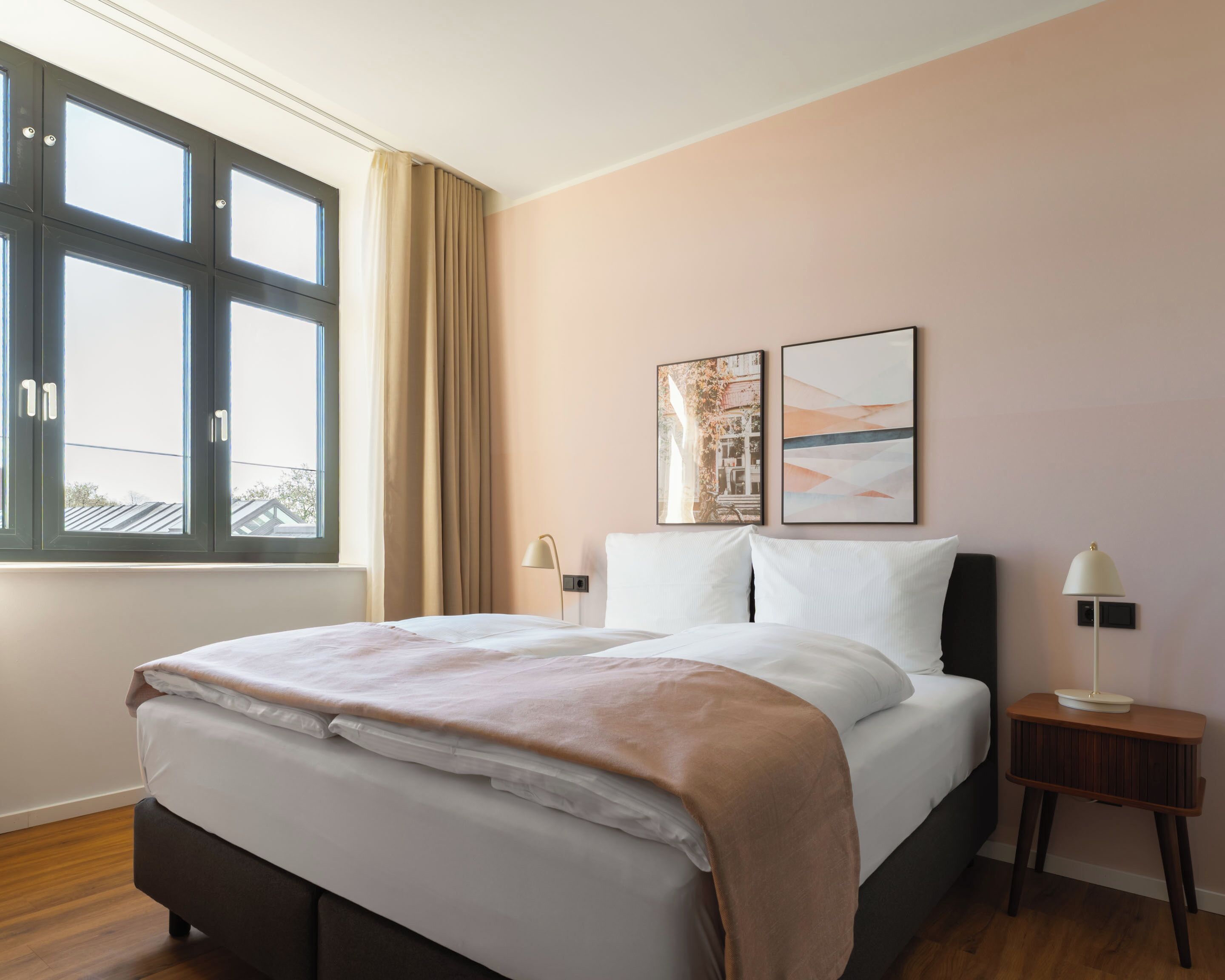 Hotelkamer met groot bed, raam met gordijnen, nachtkastje met lamp en twee kunstwerken aan de muur.