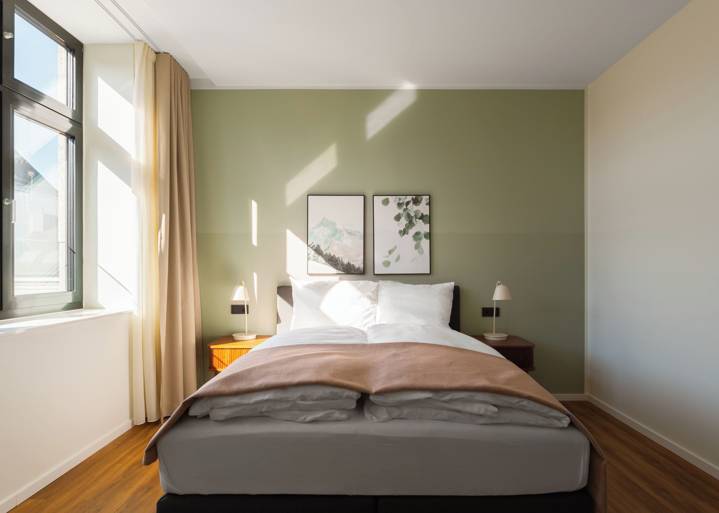 A neatly made bed in a room with a large window, two wall-mounted picture frames, and bedside lamps.