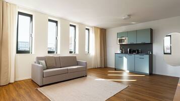 A modern living room with a sofa, a kitchen area with appliances, and large windows.