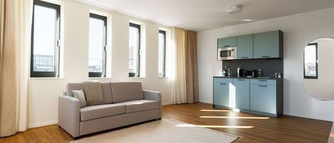 Una sala de estar moderna con un sofá, una cocina equipada y ventanas amplias.
