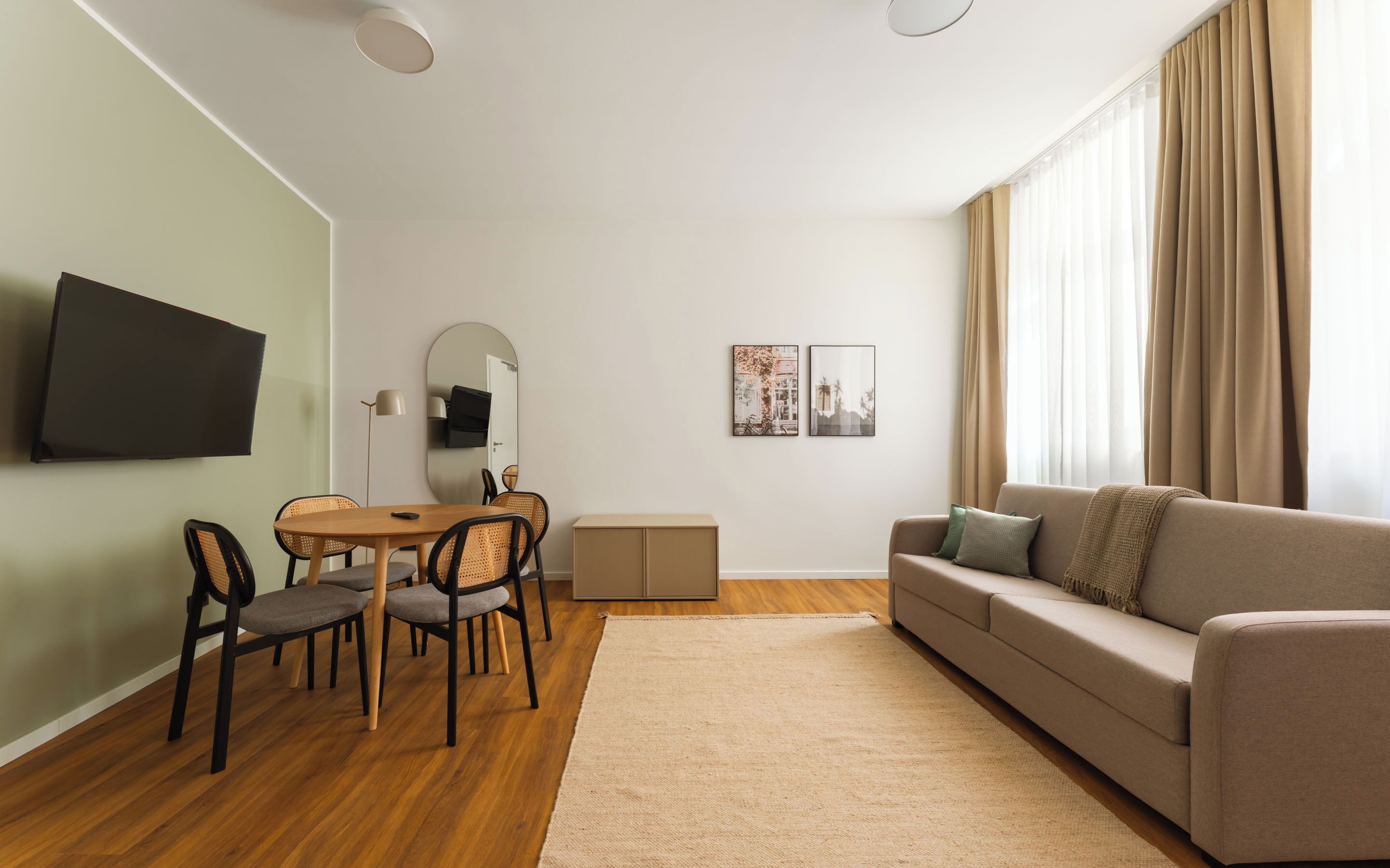 A living room with a sofa, a dining table with chairs, a flat-screen TV mounted on the wall, and a large window with curtains.