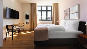 Habitación de hotel con cama, escritorio con silla, televisor montado en la pared y ventana con cortinas.