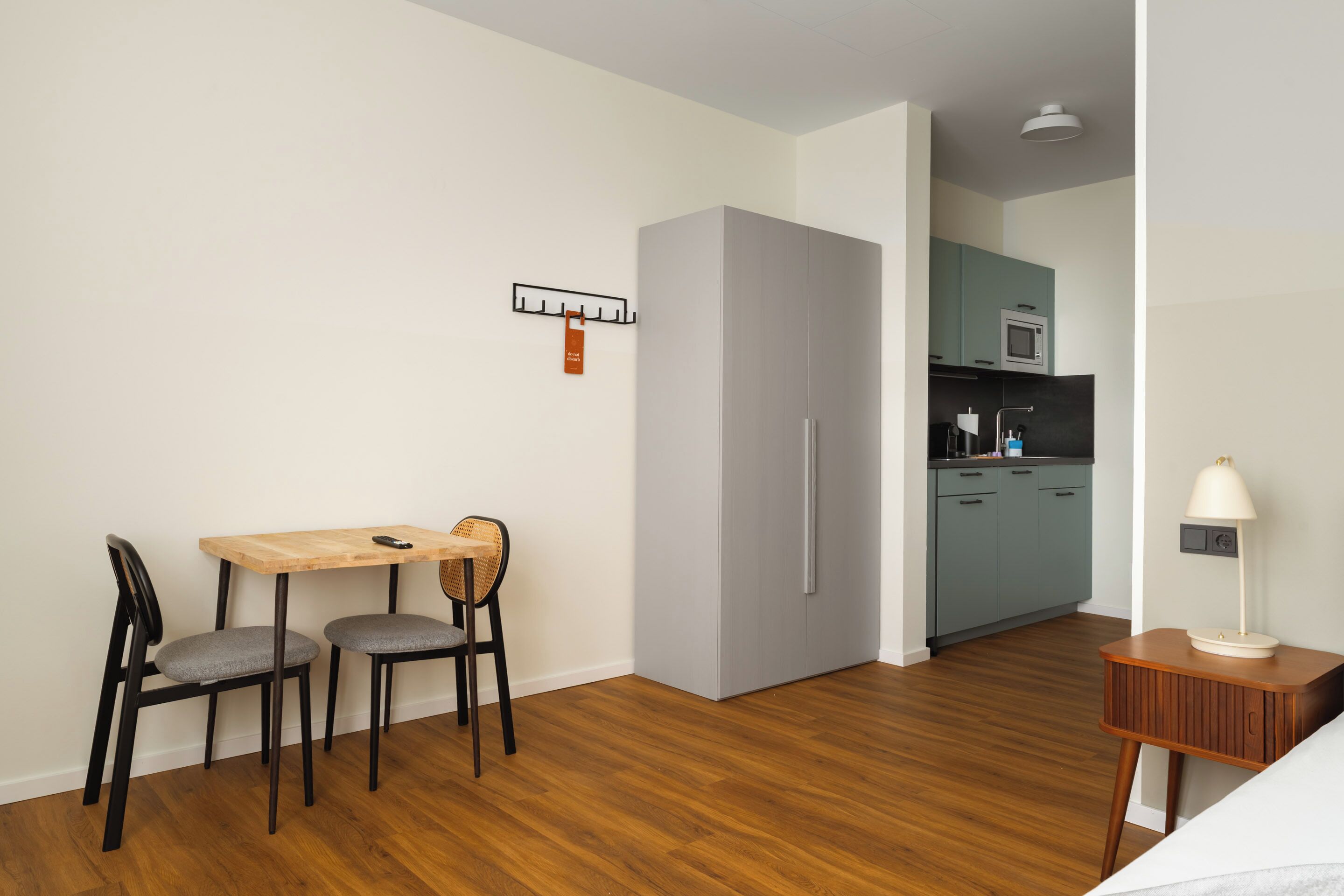 A modern hotel room with a wooden dining table, two chairs, a small side table with a lamp, a built-in refrigerator, and a kitchenette with a microwave.