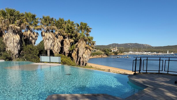 A clear swimming pool with a wooden deck, surrounded by palm trees and a view of a marina with boats. - KerTerra Corsica - Apartment 3 Bedrooms- 6 Pers - Heated pool & private beach (Pianottoli-Caldarello)