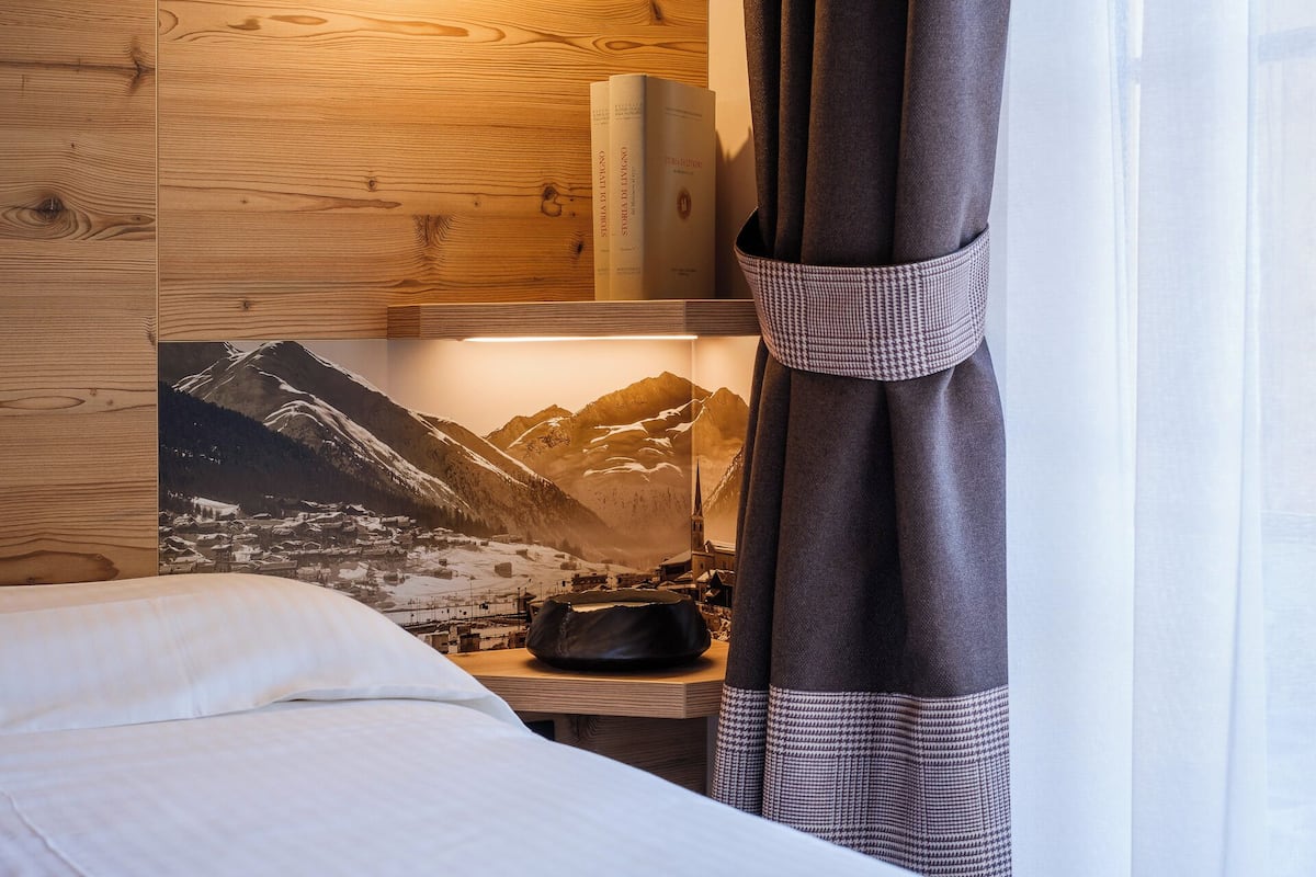 A hotel room with a bed, a wooden headboard, a nightstand with books, a lamp, and a view of mountains through a window.