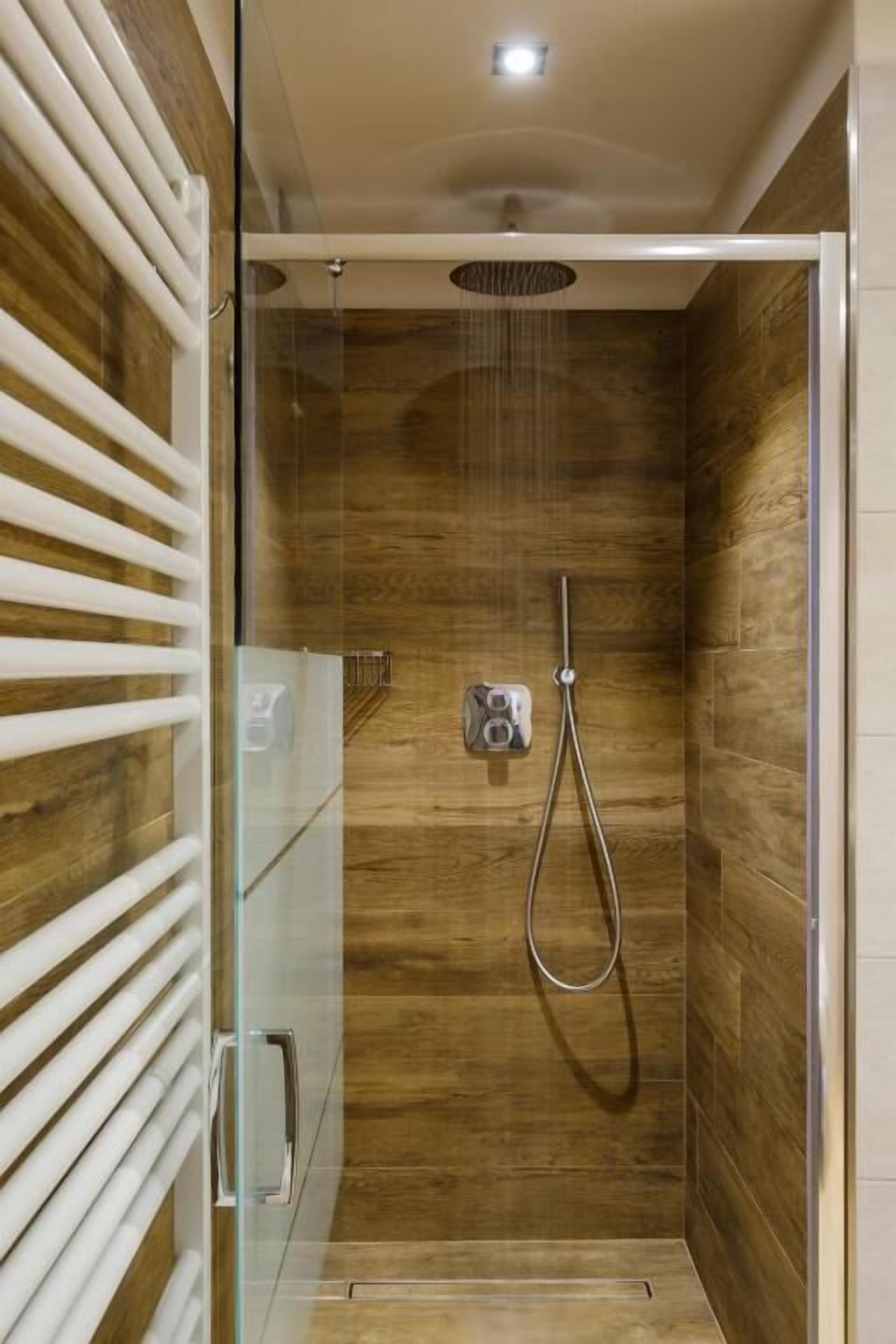 A glass-enclosed shower with a wooden wall, a handheld showerhead, and a control panel.