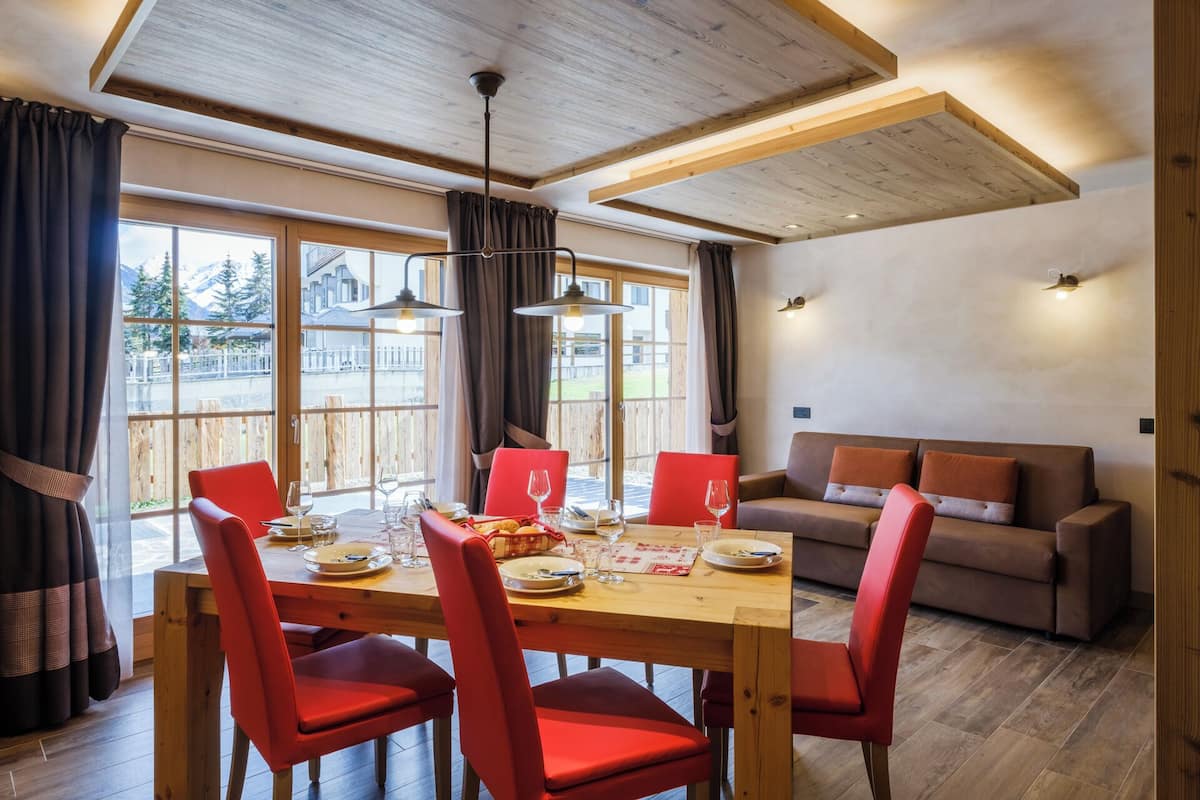 A dining area with a wooden table and red chairs, a sofa, and large windows with a view of mountains.