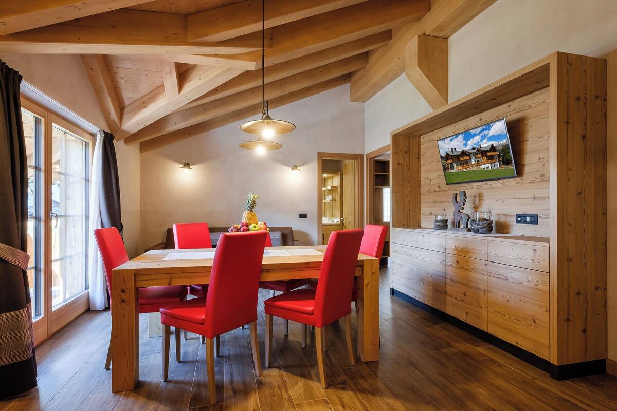 A dining area with a wooden table and red chairs, a wooden ceiling with exposed beams, a television on the wall, and a window with curtains.