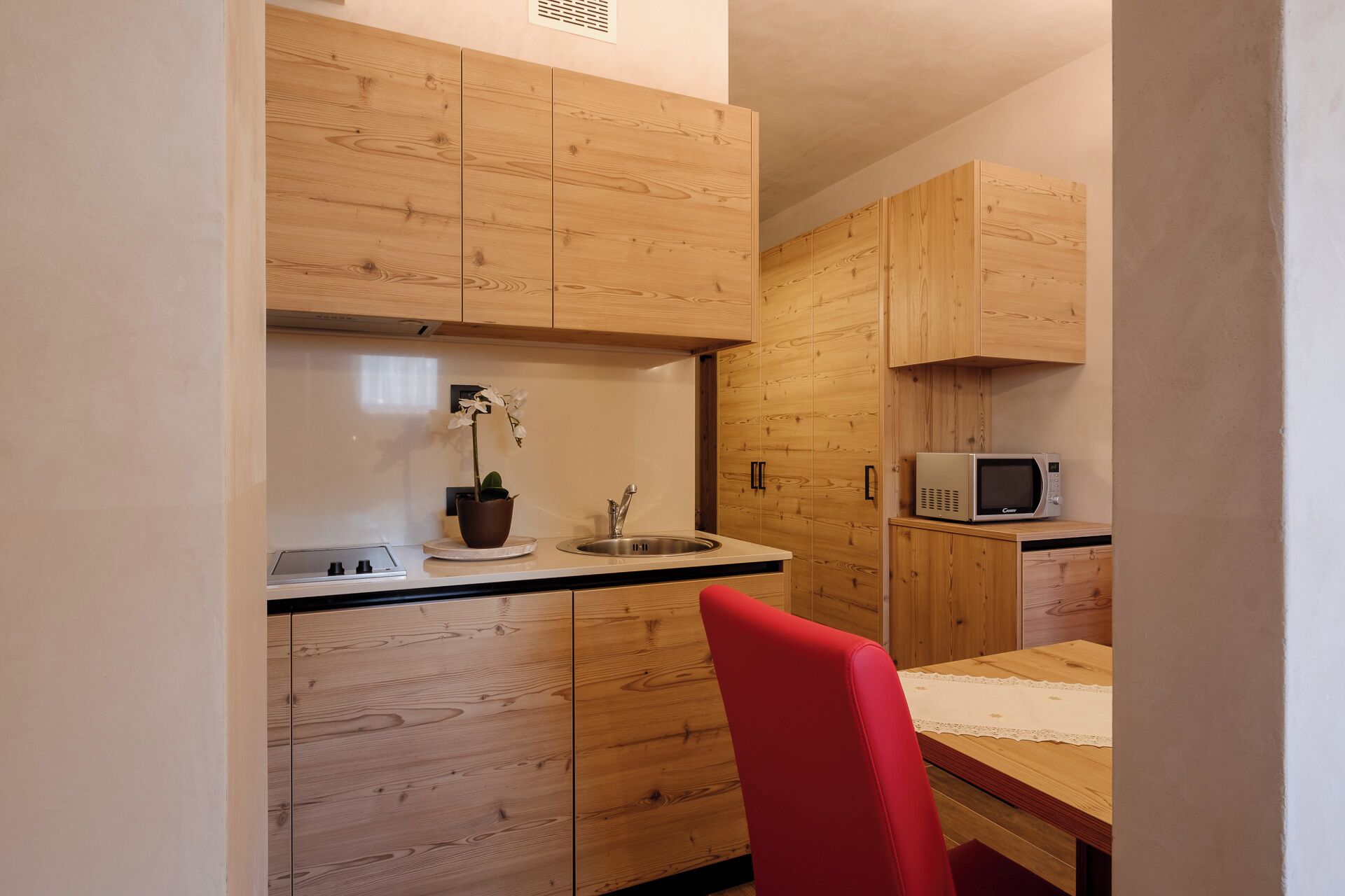 A compact kitchen with wooden cabinets, a microwave, a sink, and a red chair.