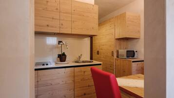 A compact kitchen with wooden cabinets, a microwave, a sink, and a red chair.