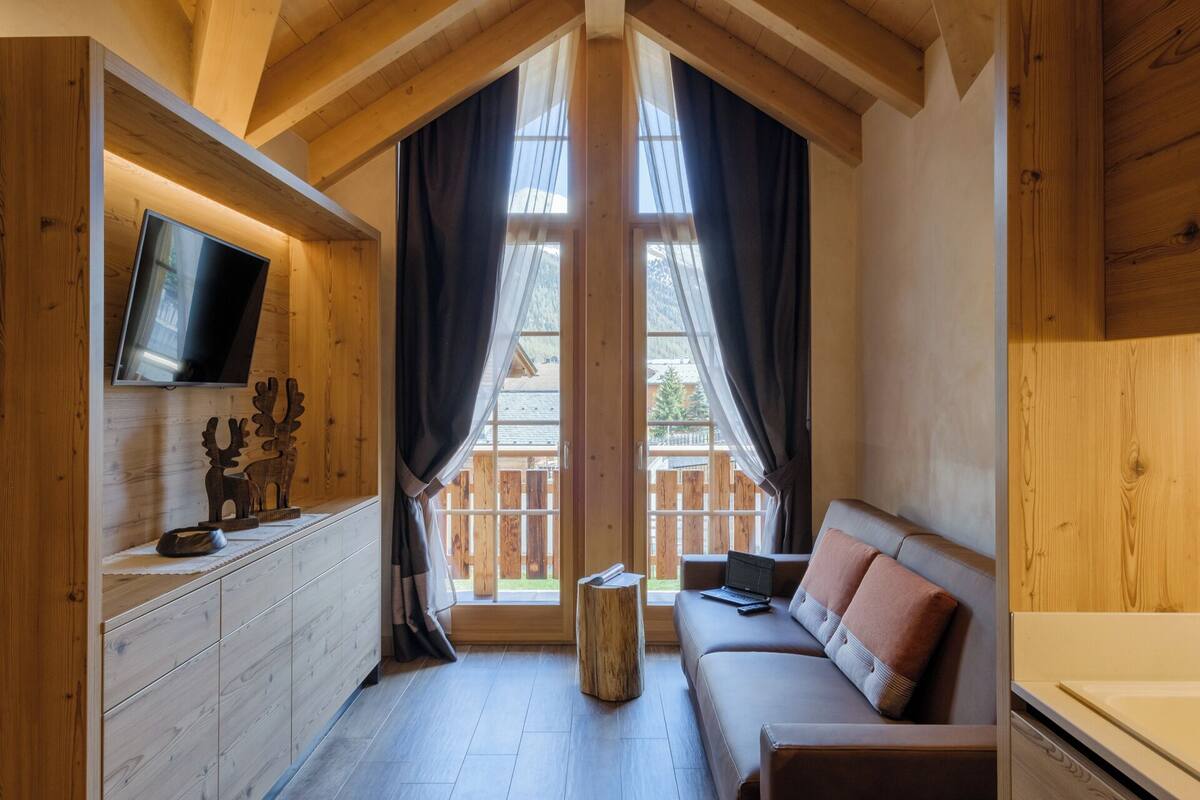 A cozy cabin interior with a wooden ceiling, a mounted TV, a sofa with cushions, a small wooden stool, and a window with curtains.