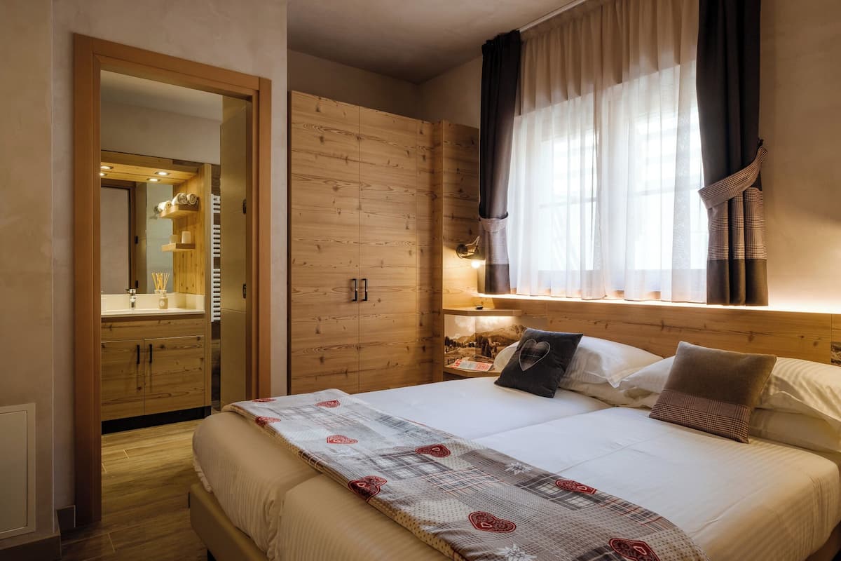 A bedroom with a wooden wardrobe, a bed with a patterned blanket, a nightstand with a lamp, and a window with sheer curtains.