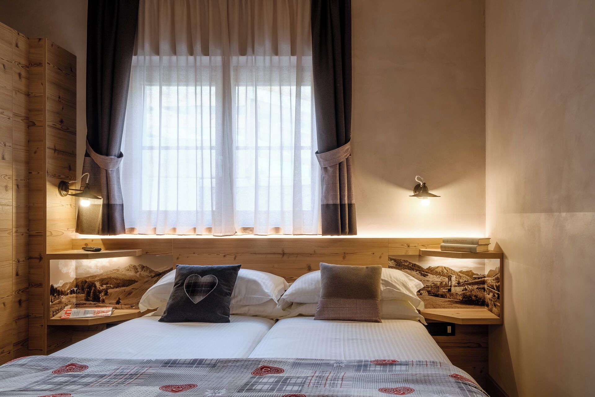 Un dormitorio con una cama, dos mesitas de noche, un ventanal con cortinas translúcidas, una lámpara de pared y un cartel en la pared.